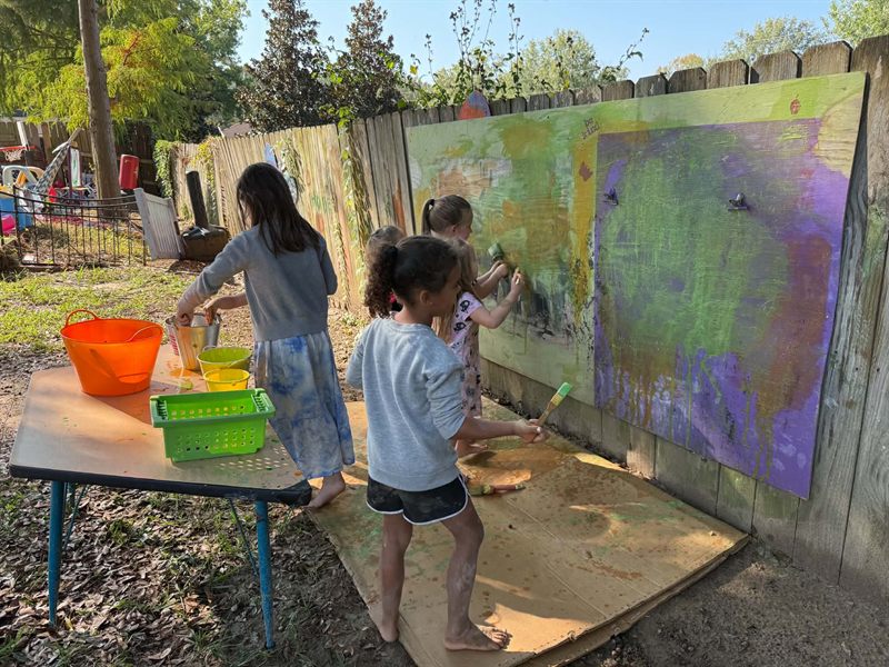 Children painting a large mural on a board propped against the backyard fence