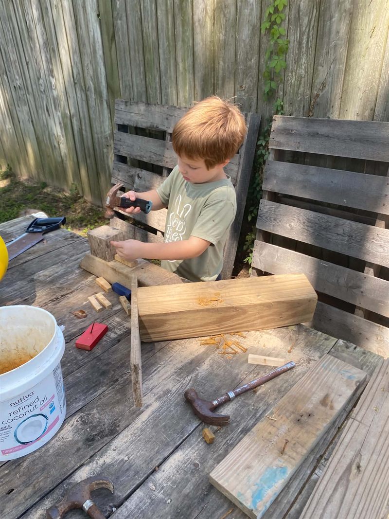 A boy hammering a nail into a block of wood at an outdoor workbench